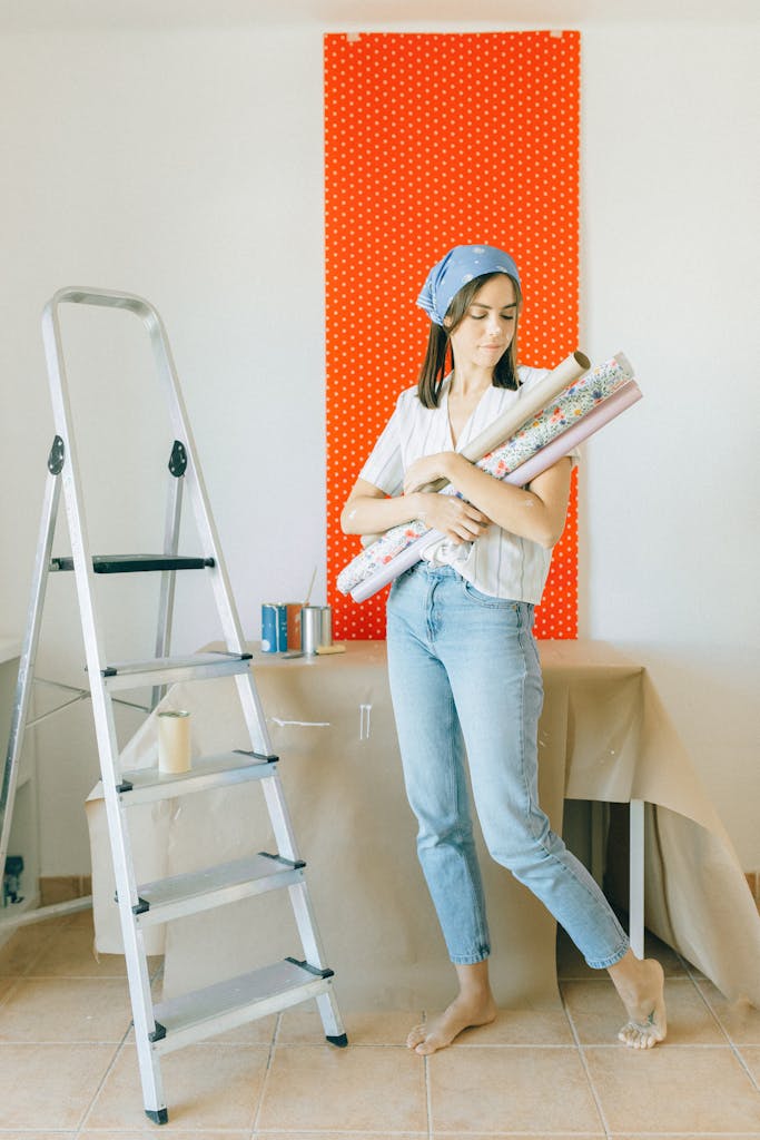 Woman holding wallpaper rolls ready to decorate a room with a ladder and tools nearby.